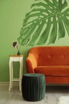 Stylish sofa with table and ottoman near green wall with drawing of tropical leaf