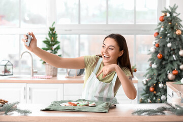 Beautiful housewife near plate with gingerbread cookies taking selfie in kitchen