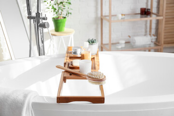 Wooden bathtub tray with different supplies and cosmetic products in light bathroom