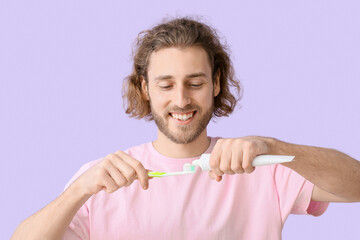 Young man applying tooth paste onto brush on lilac background