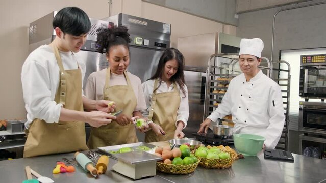 Hobby cuisine course, senior male chef in cook uniform teaches young cooking class students to peel and chop apples, ingredients for pastry foods, fruit pies in restaurant stainless steel kitchen.