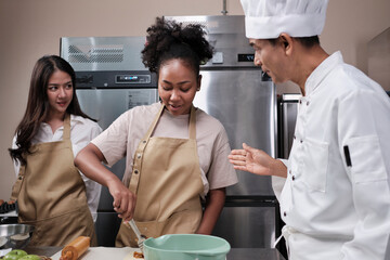 Cuisine course, senior male chef in cook uniform teaches young cooking class students to knead and roll pastry dough, prepare ingredients for bakery foods, fruit pies in stainless steel kitchen.