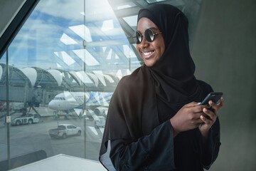 Portrait of smiling African Muslim woman in traditional black hijab, wearing sun glasses and using...