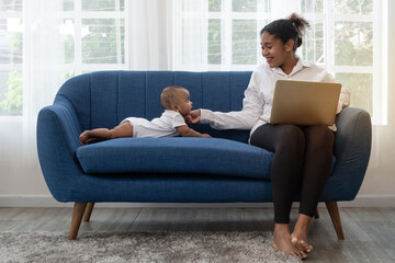 African mother working remotely on laptop while taking care of her baby on couch, freelance single mom working at home