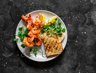Grilled shrimp skewers with bread, lemon and micro greenery on a dark background, top view