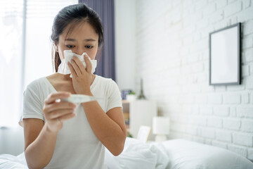 Sick day at home. Young asian woman lying on the bed have a fever and measure body temperature. Home isolation concept.