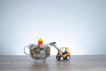 A glass cup filled with coins and a house model on the top. A toy loader loading the coins. Property value concept 