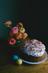 A vase with ranunculus flowers on the table along with Easter bread and colored eggs