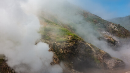 Clouds of steam from hot springs envelop the mountain slopes.  Green vegetation on the hills. Blue sky. Poor visibility due to haze. Kamchatka. Valley of Geysers