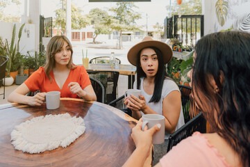 Three hispanic young adult women gossiping at a cafe, surprised and shocked. Holding a mug on their...