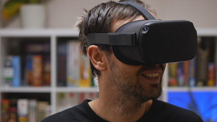 A bearded man wearing VR glasses smiles at the virtual world he sees. Close-up. In the background is a rack of games. VR helmet for entertainment, education, and gaming.