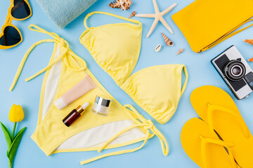 Top view of yellow bikini and cream bottle, sandals, sunglasses with beach accessories on the blue background. Summer time concept.