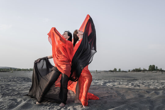 Two Girls In Black And Red Flowing Fabric Pose On Sand Dune