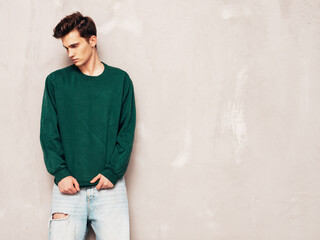 Portrait of handsome confident stylish hipster lambersexual model.Man dressed in green sweater and jeans. Fashion male posing in studio near grey wall