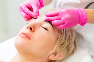 Obraz na płótnie Canvas Mesotherapy and rejuvenation. Portrait of an adult blonde woman at a cosmetologist's appointment. A beautician in pink medical gloves applies a needle to the client's face