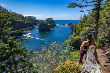 Adventurous athletic woman standing on a cliff edge at Cape Flattery looking at the beautiful Pacific Northwest coast on a sunny day.