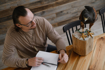 Man is working, writing with wild bird at home by the table. Making noted, memories, diary with...