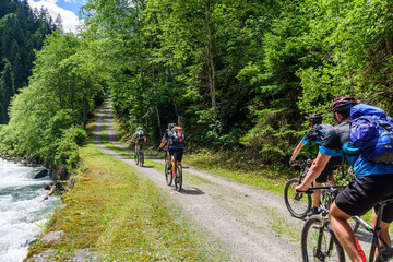 Fototapeta premium Mehrere Mountainbiker fahren an einem Wildbach in den Alpen entlang 