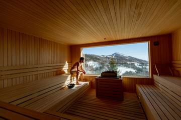 Young man relaxing in the sauna and watching winter forest through the window