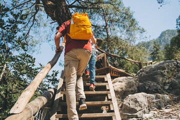 Rear view young adult man climbing a wooden ladder in the mountains. Casually dressed tourist with yellow backpack going up the wooden crafted ladder in the national park with big boulders and cliffs.