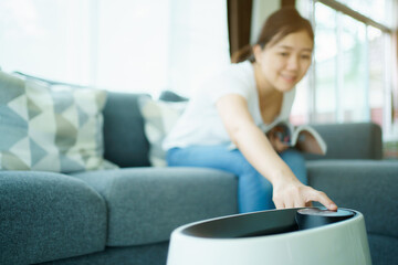 Happy Asian young woman turning on high efficiency air purifier while staying and reading a book in...
