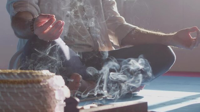Man sitting in lotus pose with smoke from incense around him. Medium shot of man doing yoga in studio, meditating, recovering strength, enjoying fragrance of burning sticks. Meditation, smoke concept
