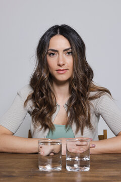 Young woman with glasses of water sitting at table against gray background