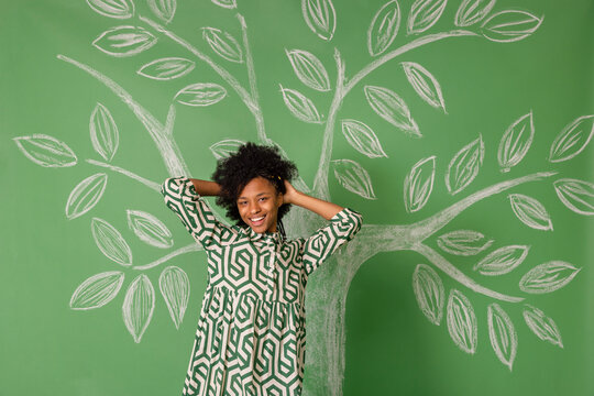 Happy young woman with hand in hair standing in front of tree drawing on green wall
