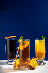 Collection of autumn winter hot drinks. Set of hot lemon ginger tea, mulled wine, fruit citrous punch grog in warm cocktail glass mugs, with cinnamon, spices, dark blue white background copy space