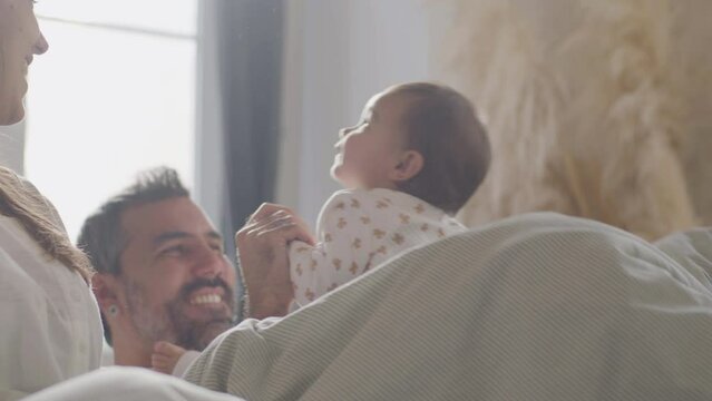 Happy married couple enjoying lazy weekend morning cuddling in bed with their baby. Father playing with daughter, swaying her back and forth on his lap and laughing. Happy family concept