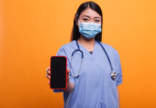 Confident Hospital Nurse Wearing Facemask And Stethoscope While Pointing Smartphone To Camera On Orange Background. Caregiver Wearing Virus Protection Mask And Medical Instrument While Holding Phone.