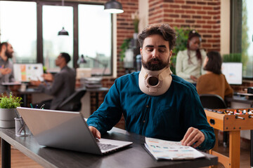 Executive manager with medical cervical neck collar while working at company turnover analyzing marketing strategy in brick wall startup office. Injured entrepreneur man planning business