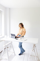 pretty blonde woman with headset stands at high table and wears high waist pants and talks with customers via notebook in a modern white office © epiximages