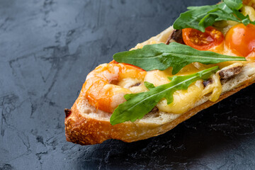Pizza cooked on a baguette with cheese, tomatoes, shrimps and arugula on a dark table, close-up.