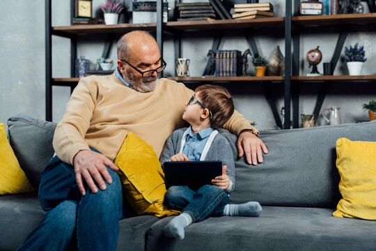 Boy with tablet PC sitting by grandfather on sofa at home