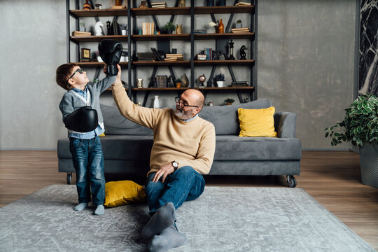 Cute boy wearing boxing gloves standing by grandfather in living room at home