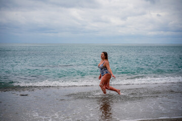 A plump woman in a bathing suit enters the water during the surf. Alone on the beach, Gray sky in the clouds, swimming in winter.