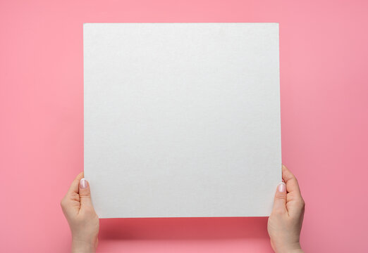 Overhead shot of woman hands holding square blank paper sheet on pink table. Woman hands with mockup empty paper sheet on pink background. Hands holding blank advertising card