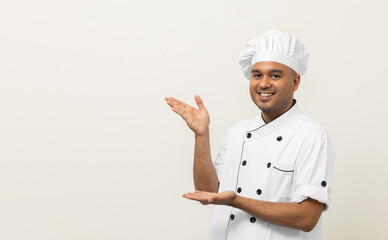 Smiling Young handsome asian man chef in uniform with hat standing showing hand palm up holding some menu on isolated. Cooking indian man Occupation chef People in kitchen restaurant and hotel.