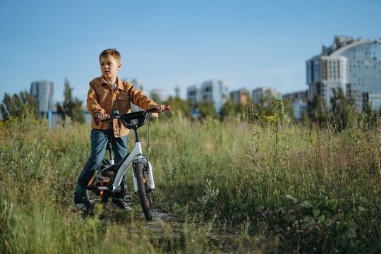 cute caucasian boy in striped henley and brown jean jacket smiling cycling on the glade of fireweed. City buildings on background. Image with selective focus