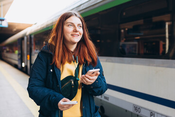 Young redhhead woman waiting on station platform with backpack on background electric train using smart phone. Railroad transport concept, Traveler