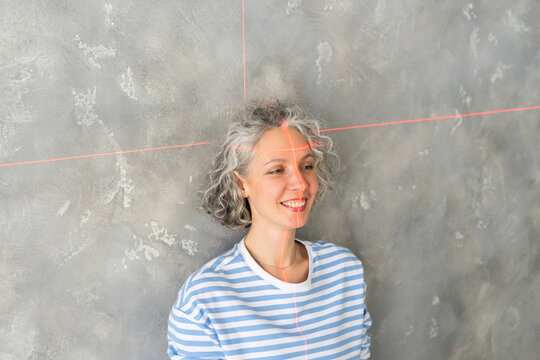 Smiling woman with laser level on face in front of gray wall