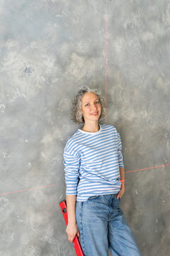 Smiling woman holding level leaning on gray wall