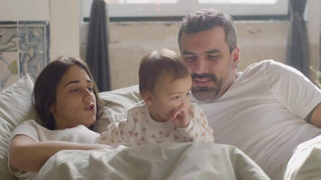 Relaxed young couple lying in bed on Sunday morning and cuddling with their cute baby girl. Young family enjoying lazy weekend while playing with cute infant and smiling. Family, parenthood concept