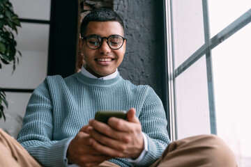 Happy man text messaging through smart phone sitting by window at home