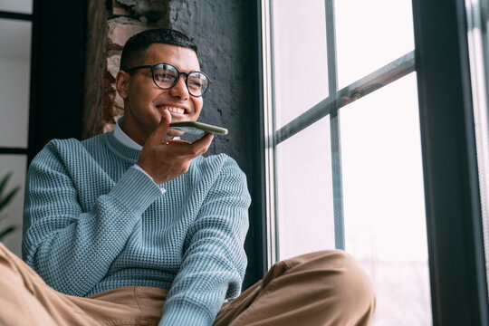 Happy young man sending voicemail through mobile phone sitting by window at home