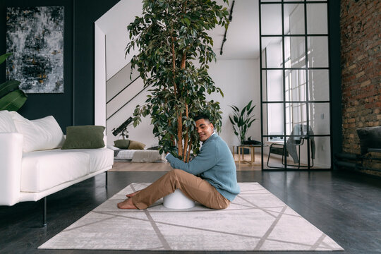 Smiling Young Man With Eyes Closed Hugging Houseplant Sitting On Carpet At Home