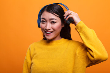 Cute joyful positive asian person stops listening to music to talk with mate while on orange background. Beautiful smiling heartily woman taking off headphones when she meets her friend.