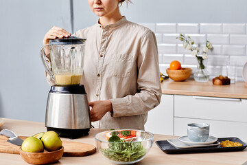 Close-up of young woman standing at kitchen table and making fresh cocktail from fruits in blender