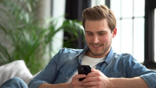 Happy Handsome Caucasian Man Using Smartphone in Cozy Living Room at Home. Man Resting on Comfortable Sofa